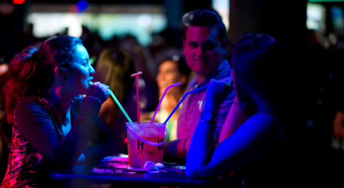 Friends gathered around a table sharing drinks at Howl at the Moon, surrounded by vibrant neon lights and a lively crowd.