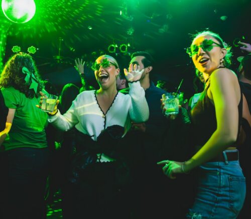 Friends dancing and celebrating at a St. Patrick’s Day party at Howl at the Moon, wearing shamrock glasses and holding drinks under green lights at a packed live music bar.