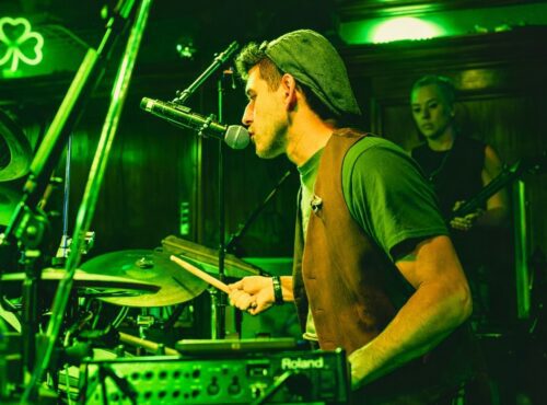 Live musician performing at Howl at the Moon during a St. Patrick’s Day party, drummer singing into a microphone under green lighting at a high-energy dueling piano bar.